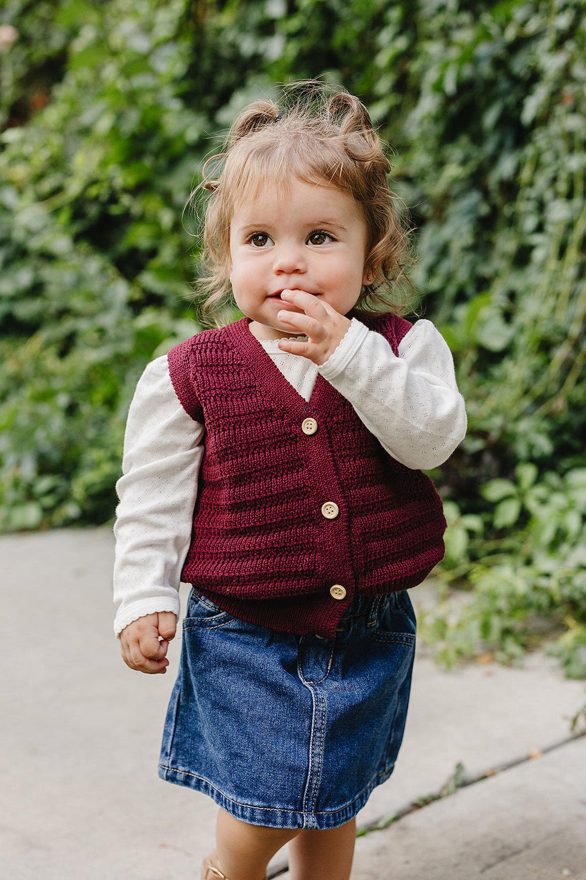 A young child is wearing a burgundy knit cotton vest over a white long-sleeve shirt, paired with a denim skirt. The backdrop features green foliage, creating a natural setting.