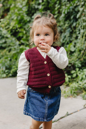 A young child is wearing a burgundy knit cotton vest over a white long-sleeve shirt, paired with a denim skirt. The backdrop features green foliage, creating a natural setting.
