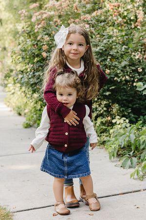 Two young girls stand on a path surrounded by greenery and flowers. The older girl wears a burgundy knit vest over a white shirt, while the younger girl is in.