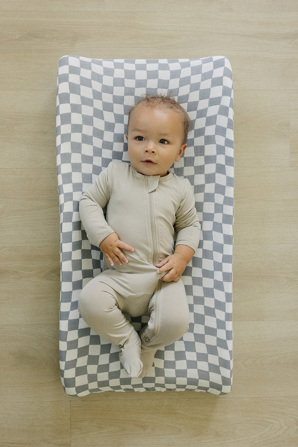 A baby in a beige onesie lies on a blue and white checkered muslin changing pad cover.