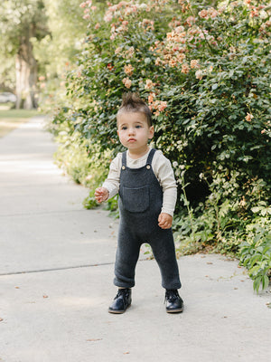 A toddler stands on a sidewalk wearing charcoal knit cotton overalls and a cream long-sleeve shirt, surrounded by lush greenery and blooming flowers, exuding a playful and stylish look.
