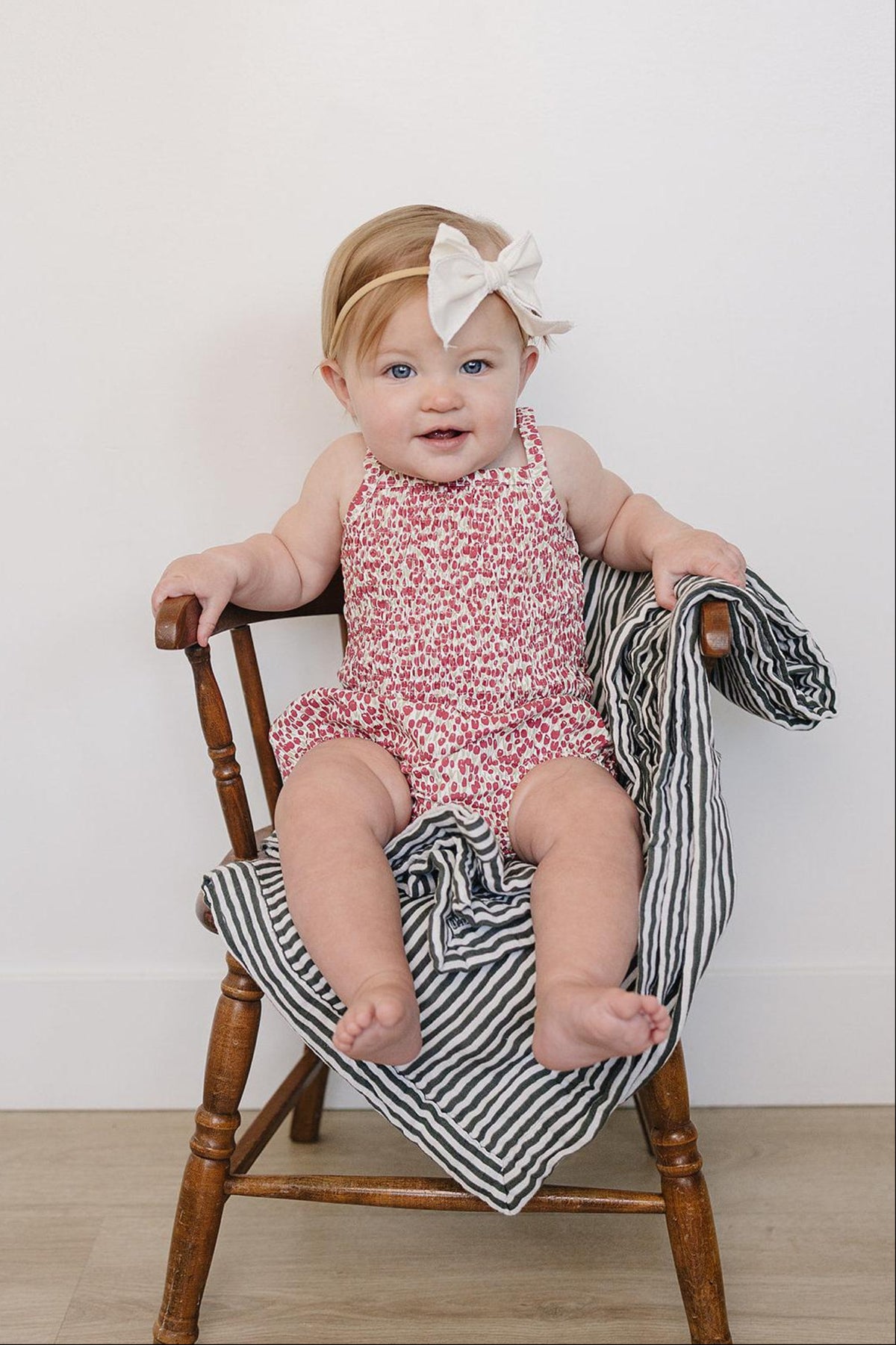 A baby girl sits on a wooden chair wearing a floral romper with a large white bow in her hair,.