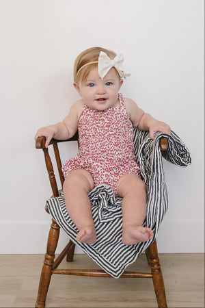 A baby girl sits on a wooden chair wearing a floral romper with a large white bow in her hair,.
