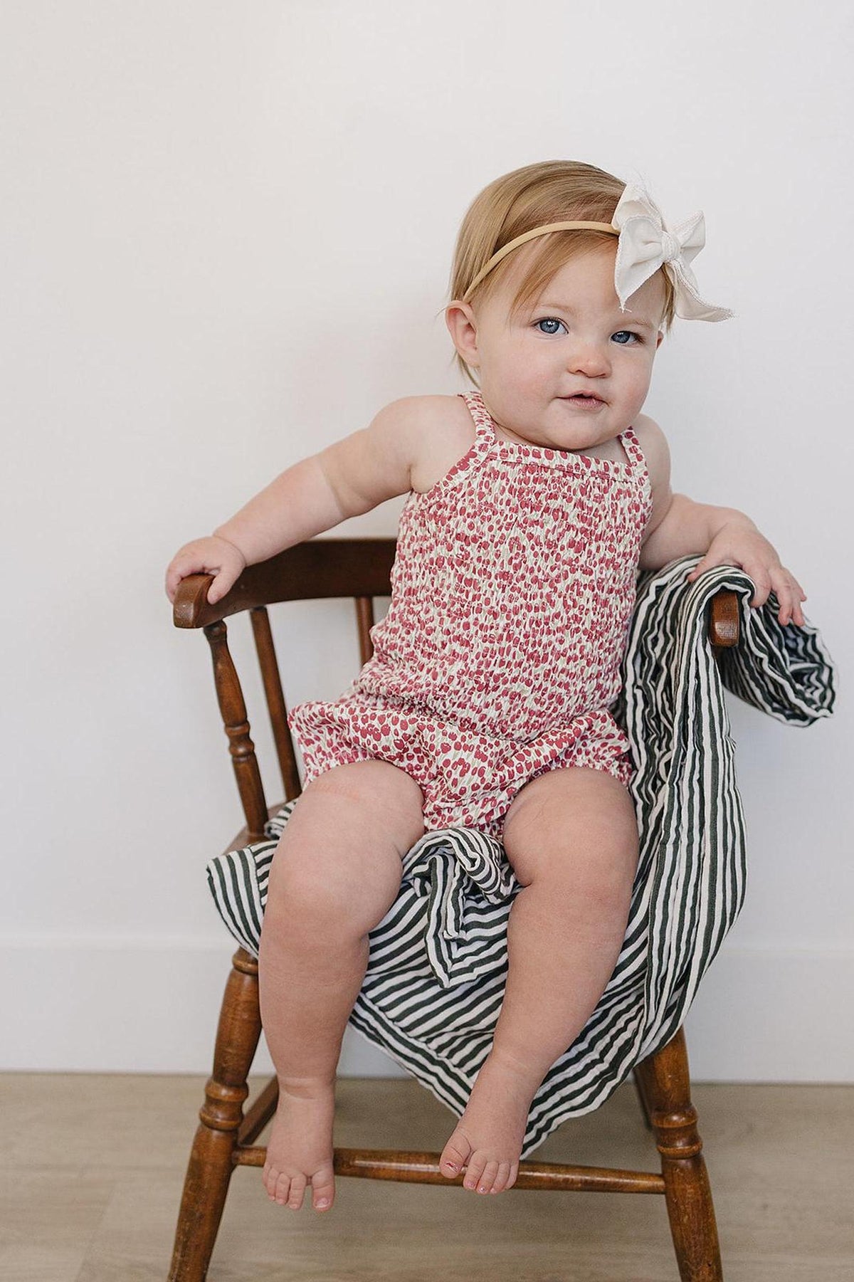 A baby girl sits on a wooden chair, wearing a floral romper and a light bow headband, with a striped.
