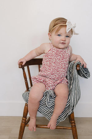 A baby girl sits on a wooden chair, wearing a floral romper and a light bow headband, with a striped.