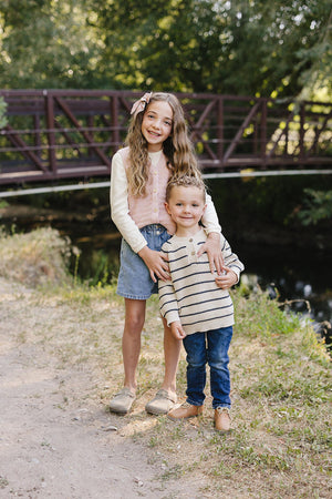 Two children smiling outdoors; one wears a pink knit vest over a white shirt, and the other a striped sweater.