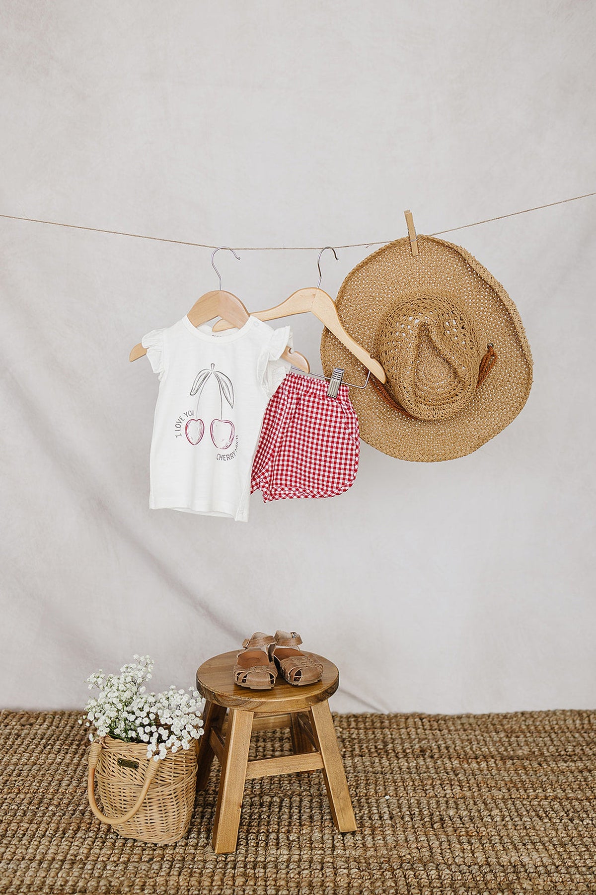 A white baby t-shirt with a cherry design and red checkered shorts hang alongside a straw hat.