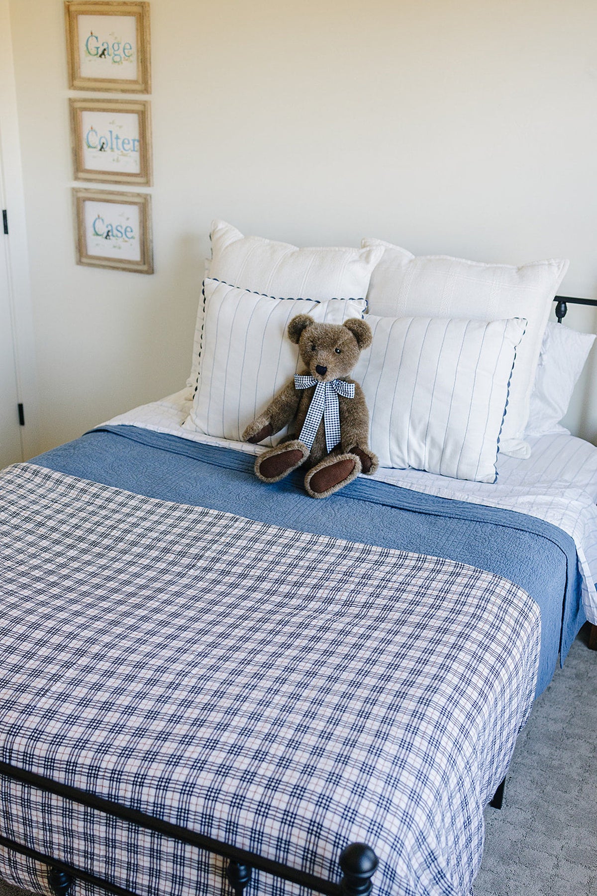 A bed with a blue and white plaid quilt, white pillows, and a teddy bear on top.