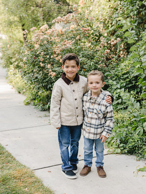Two young boys stand together on a paved path surrounded by greenery and flowering plants. One boy wears a beige cotton jacket, while the other sports a plaid flannel shirt.