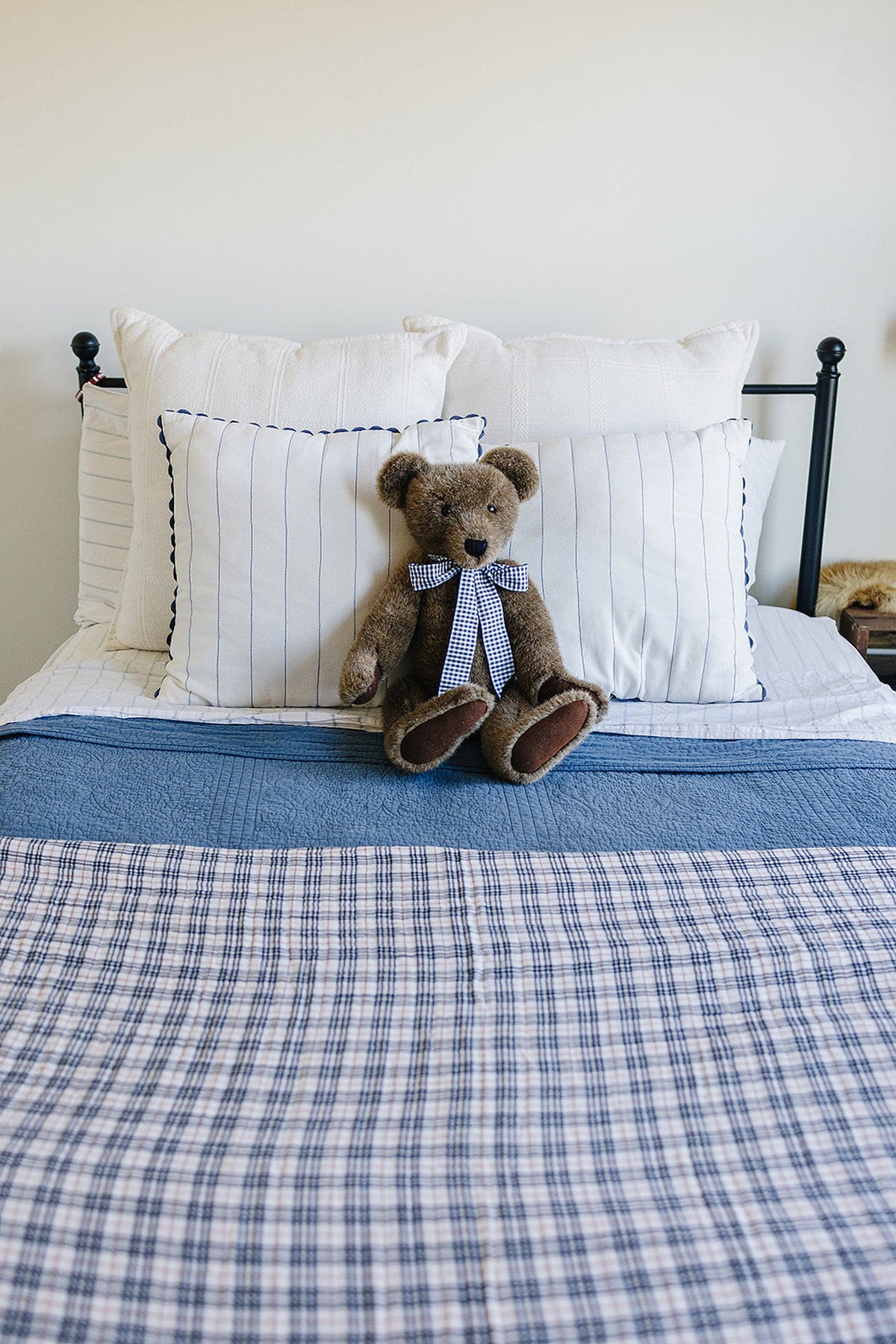 A teddy bear with a checkered bow sits on a bed with plaid and solid blue bedding.