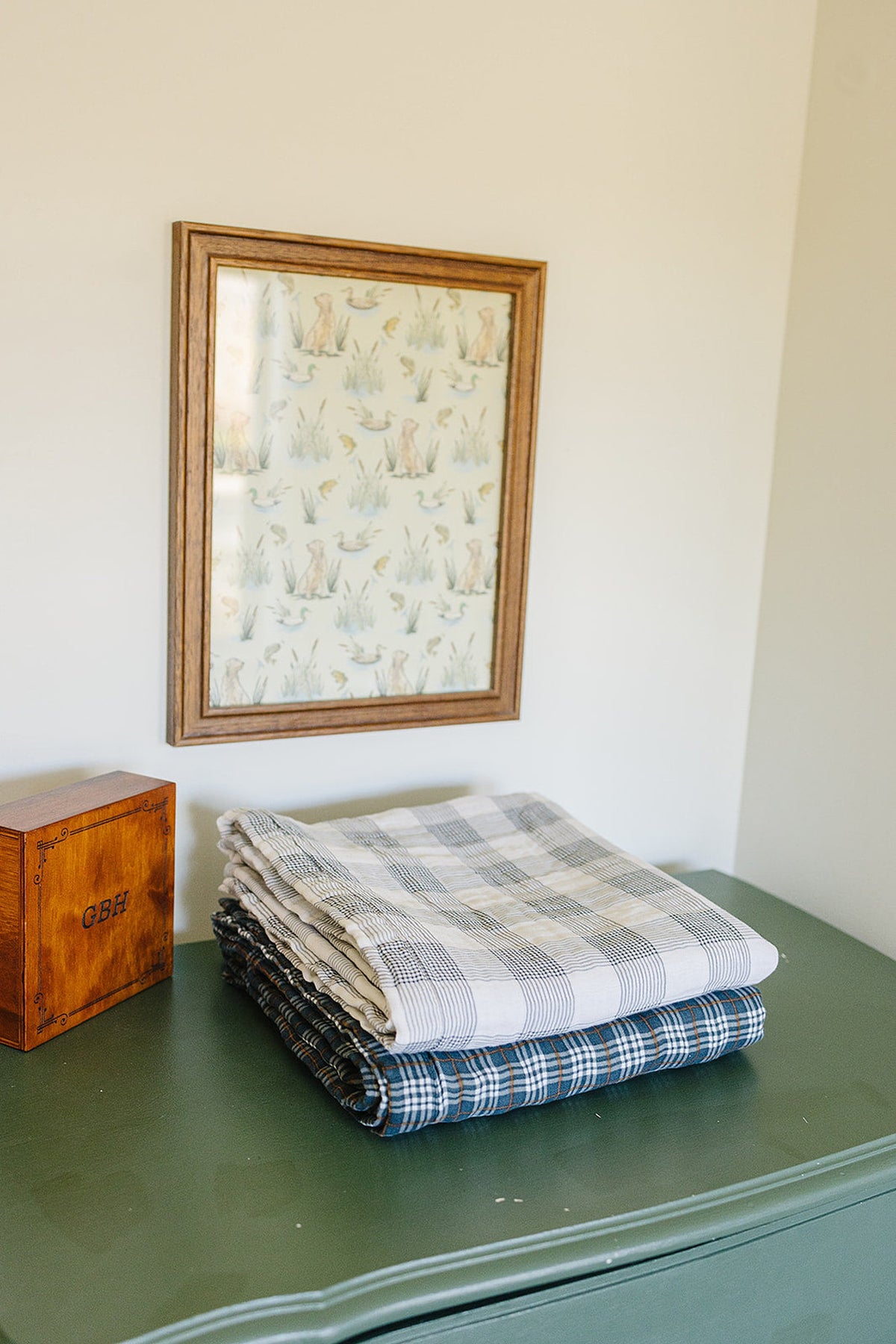 Folded plaid muslin quilts in neutral and navy tones are stacked on a green dresser beneath framed animal art.