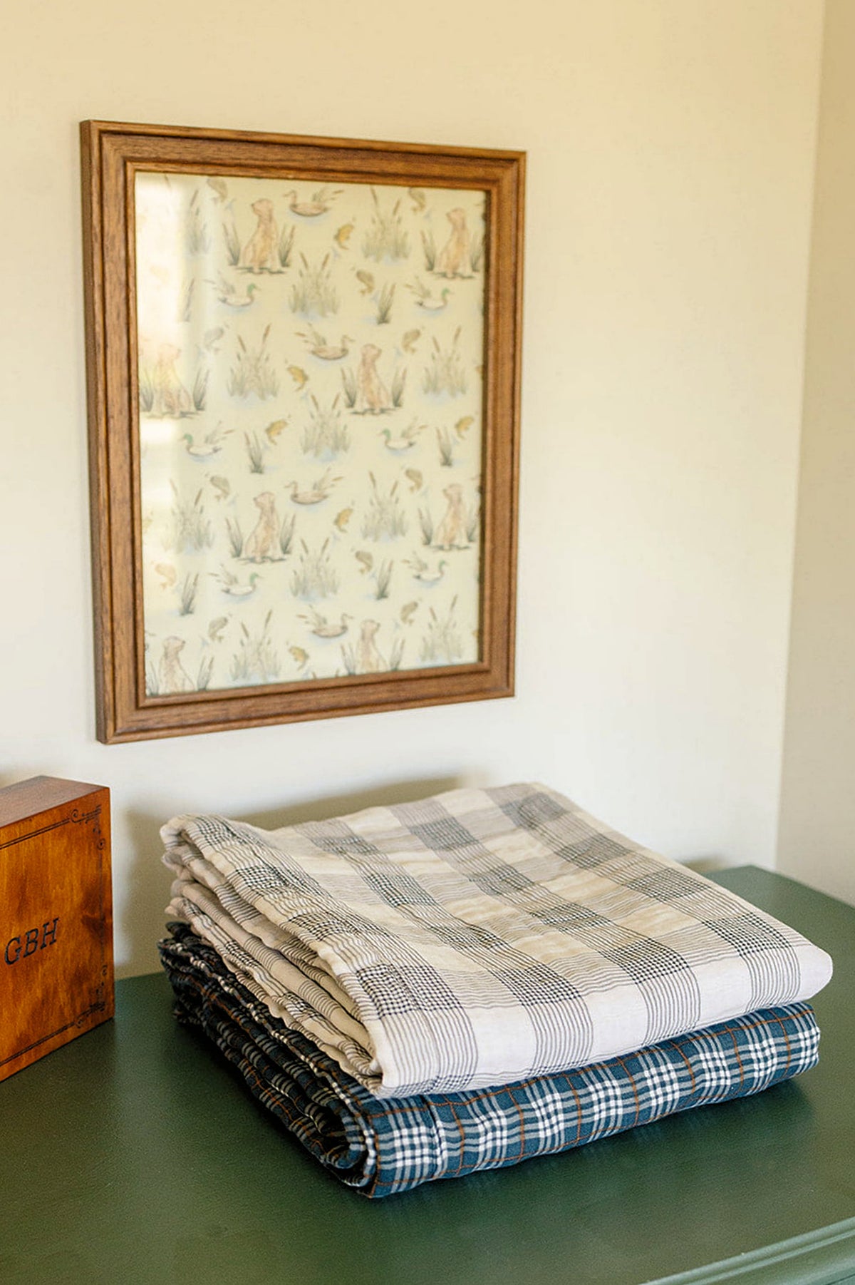 Two plaid muslin quilts, one beige and one navy, neatly folded on a green surface beneath wall art.