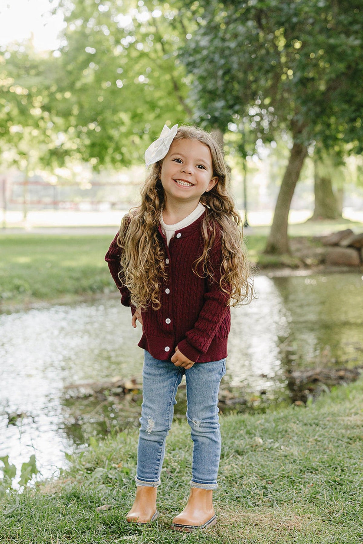 A young girl stands by a serene pond, wearing a burgundy cable knit cardigan over a white shirt and blue jeans.