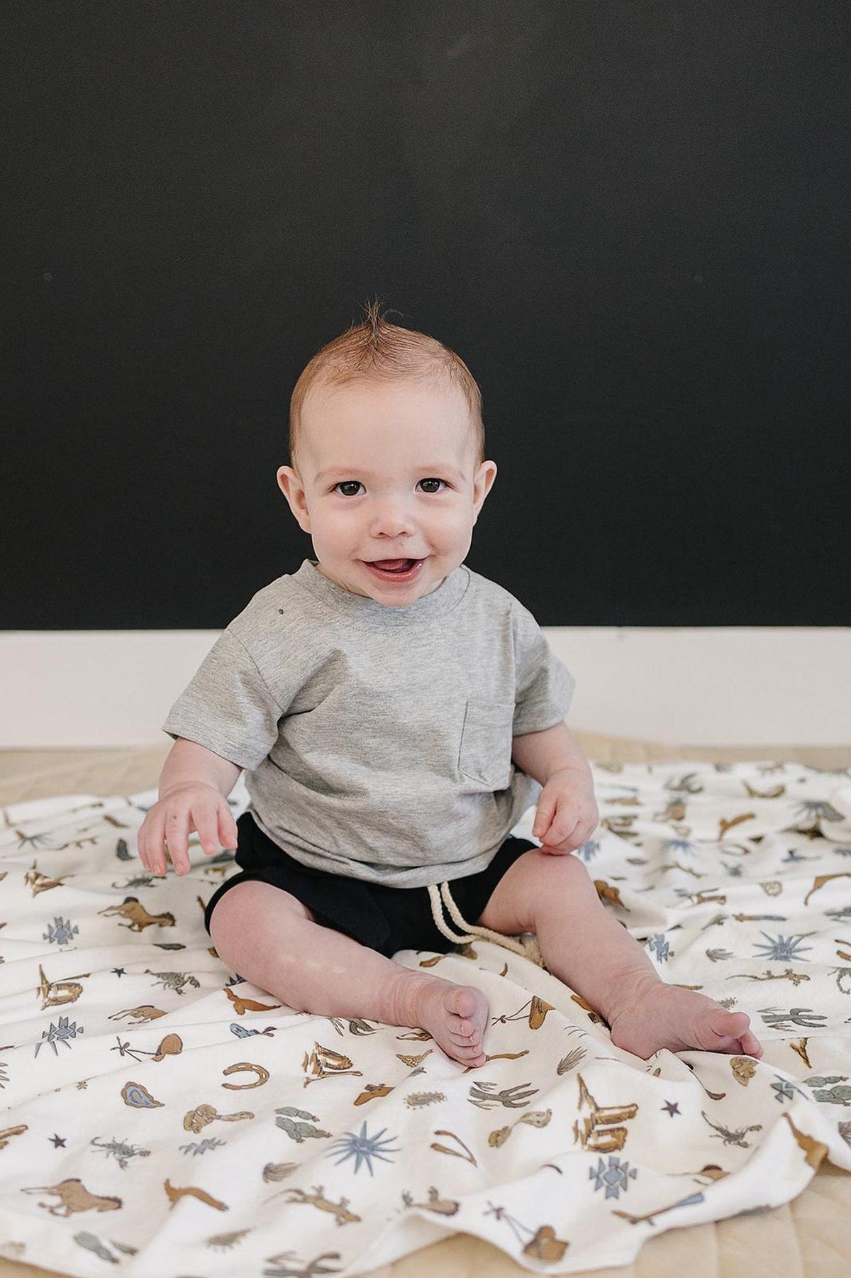 A smiling baby sits on a patterned blanket, wearing a grey cotton t-shirt with a chest pocket and black shorts.