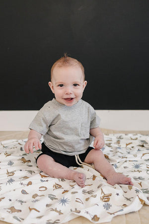 A smiling baby sits on a patterned blanket, wearing a grey cotton t-shirt with a chest pocket and black shorts.