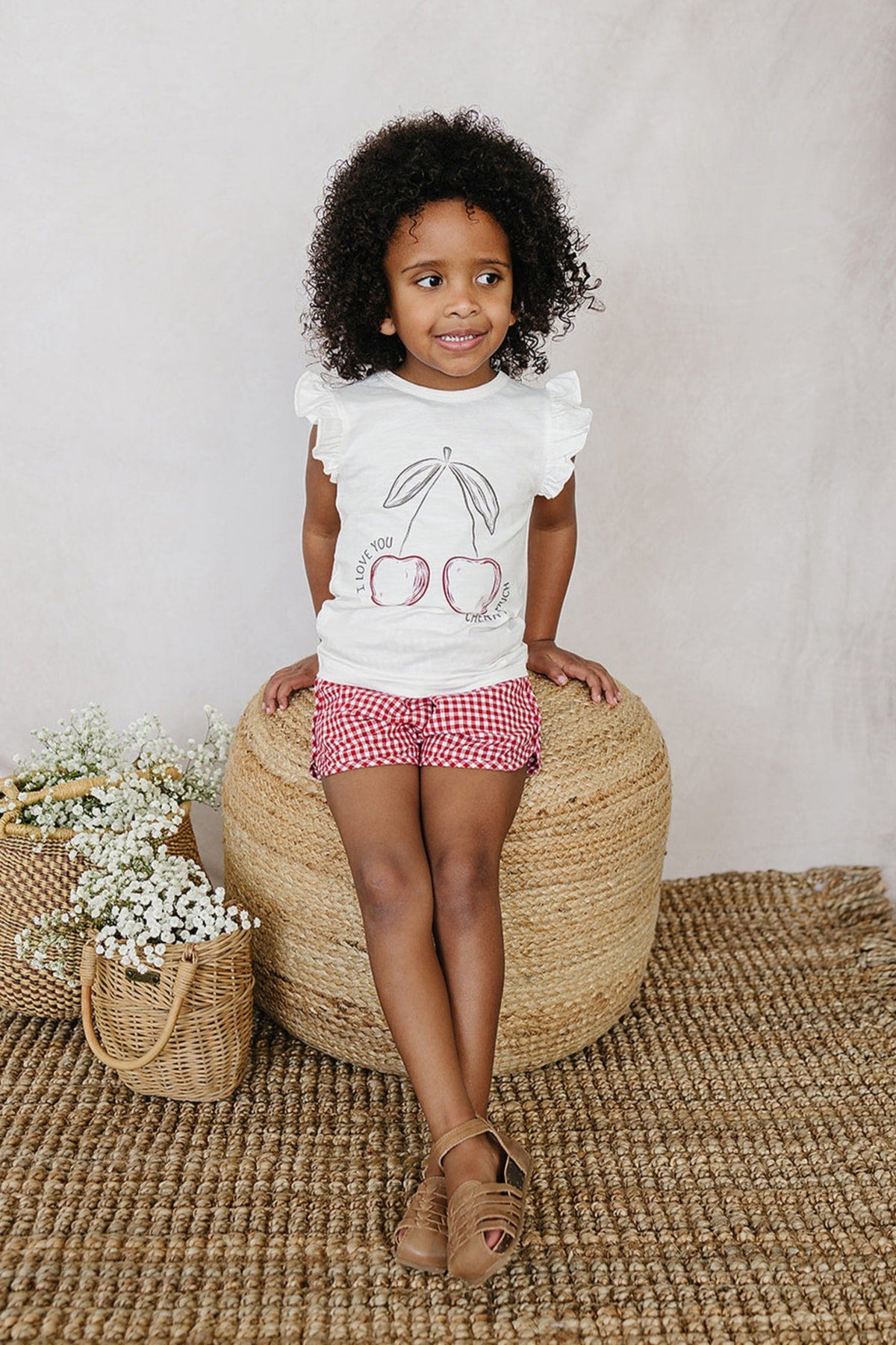 A child wearing red gingham shorts and a white cherry-print shirt sits on a woven ottoman, surrounded by baskets.