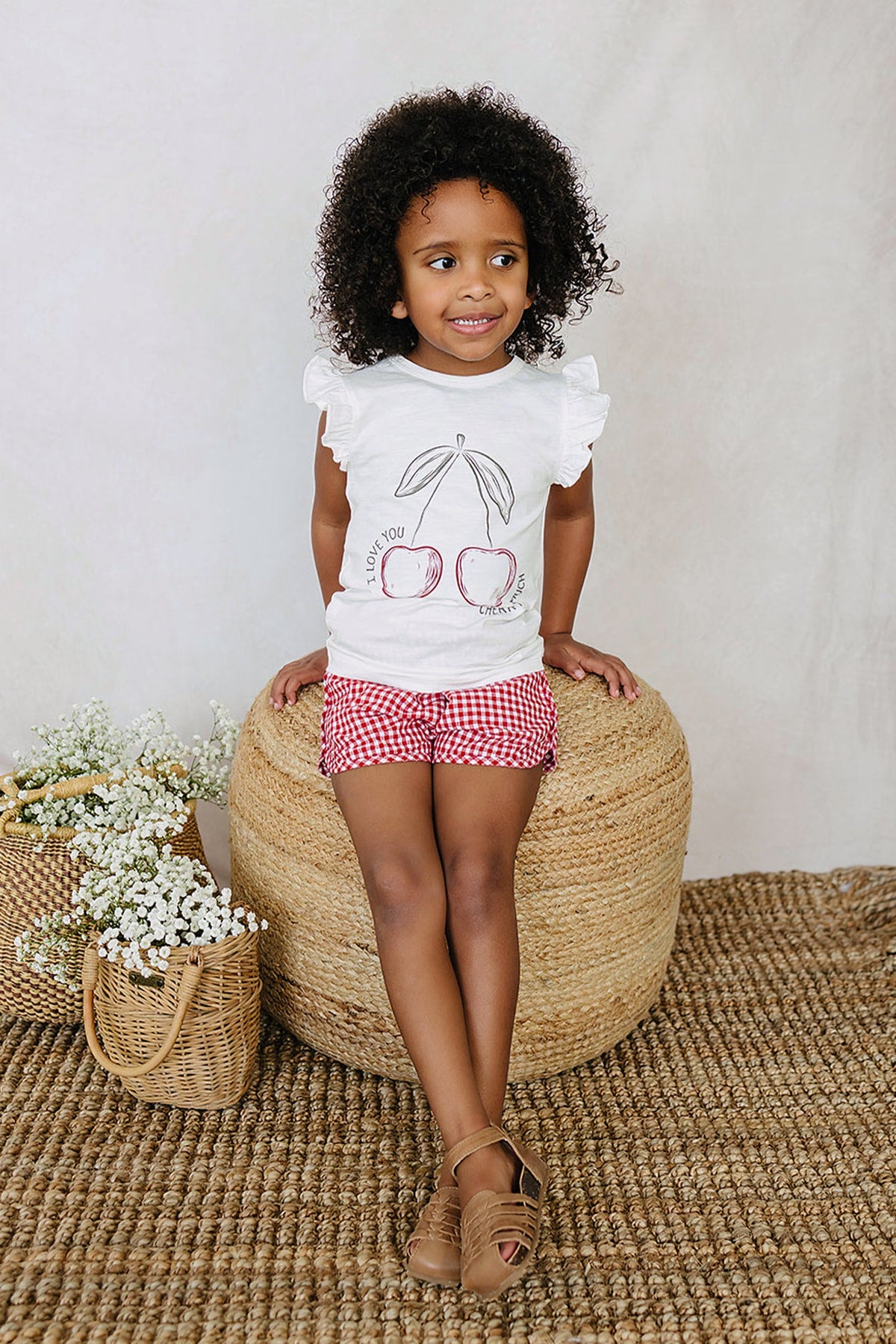 A child wearing a white ruffled t-shirt with a cherry design and red checkered shorts sits on a woven ottoman.
