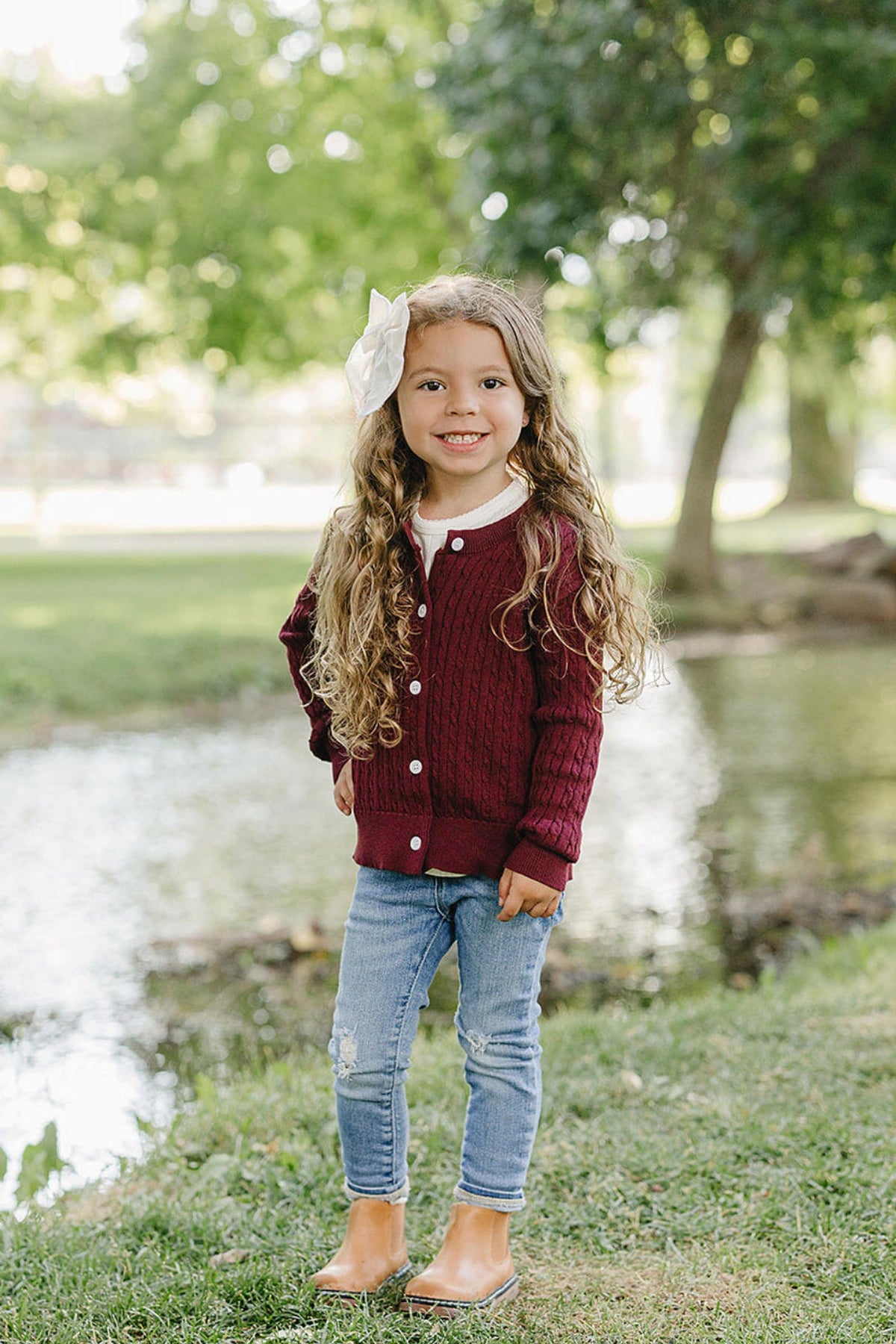 A young girl with long, curly hair stands by a pond, wearing a burgundy cable knit cardigan and light blue jeans. The natural setting features green trees and grass.