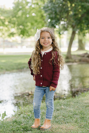 A young girl with long, curly hair stands by a pond, wearing a burgundy cable knit cardigan and light blue jeans. The natural setting features green trees and grass.