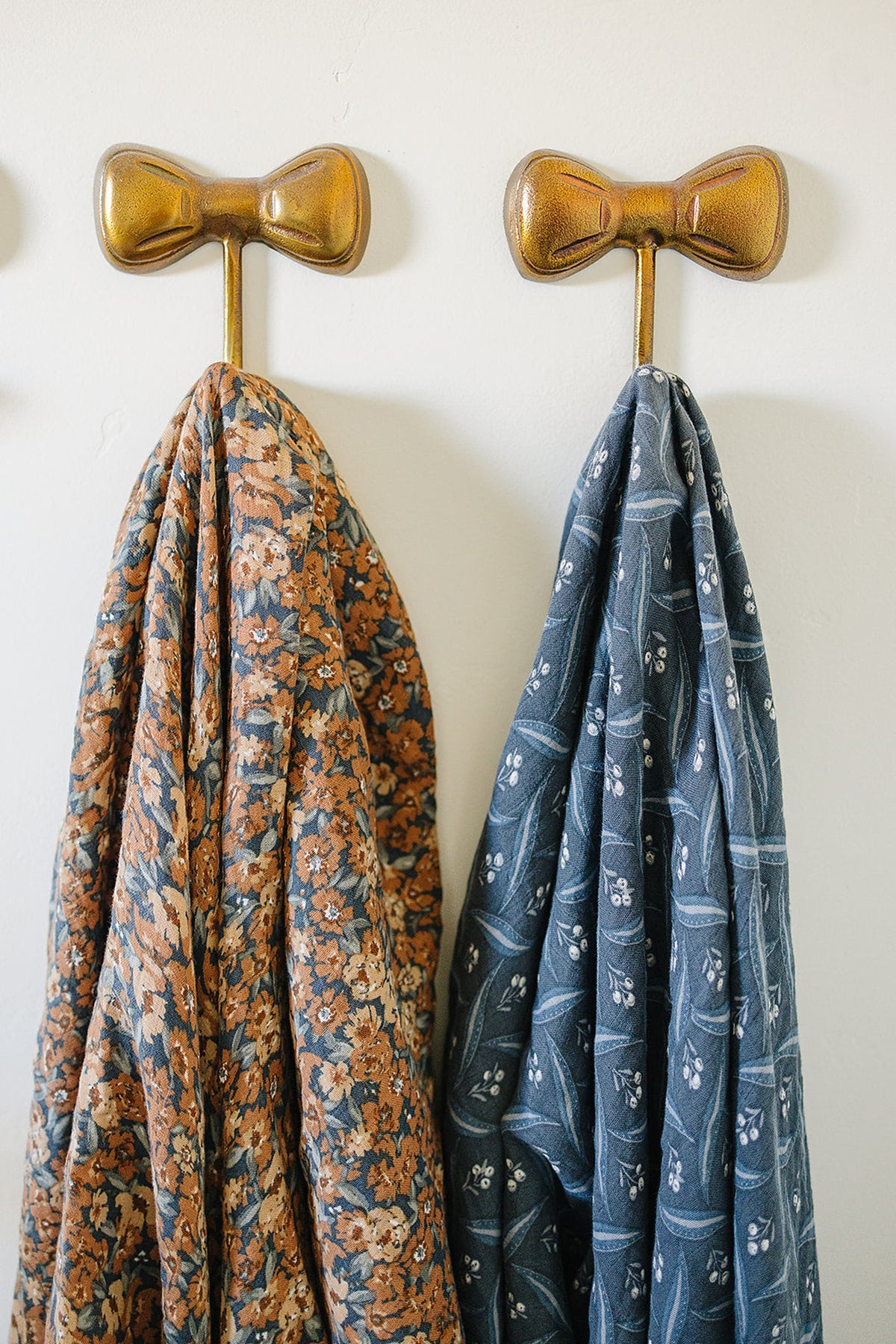 Two patterned muslin blankets hang on gold bow-shaped hooks; one is floral, the other navy with berries.