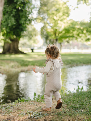 A toddler in taupe knit overalls with ruffle details walks playfully near a serene pond surrounded by lush greenery. The soft cotton fabric adds comfort to her charming outdoor outfit.