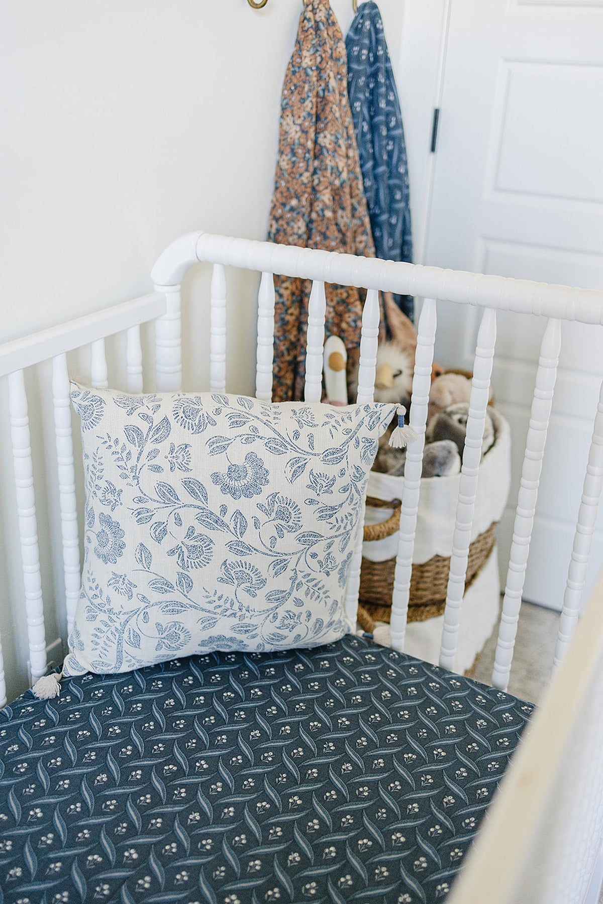 White crib with a navy patterned sheet and floral pillow, adjacent to hanging blankets and a woven basket.