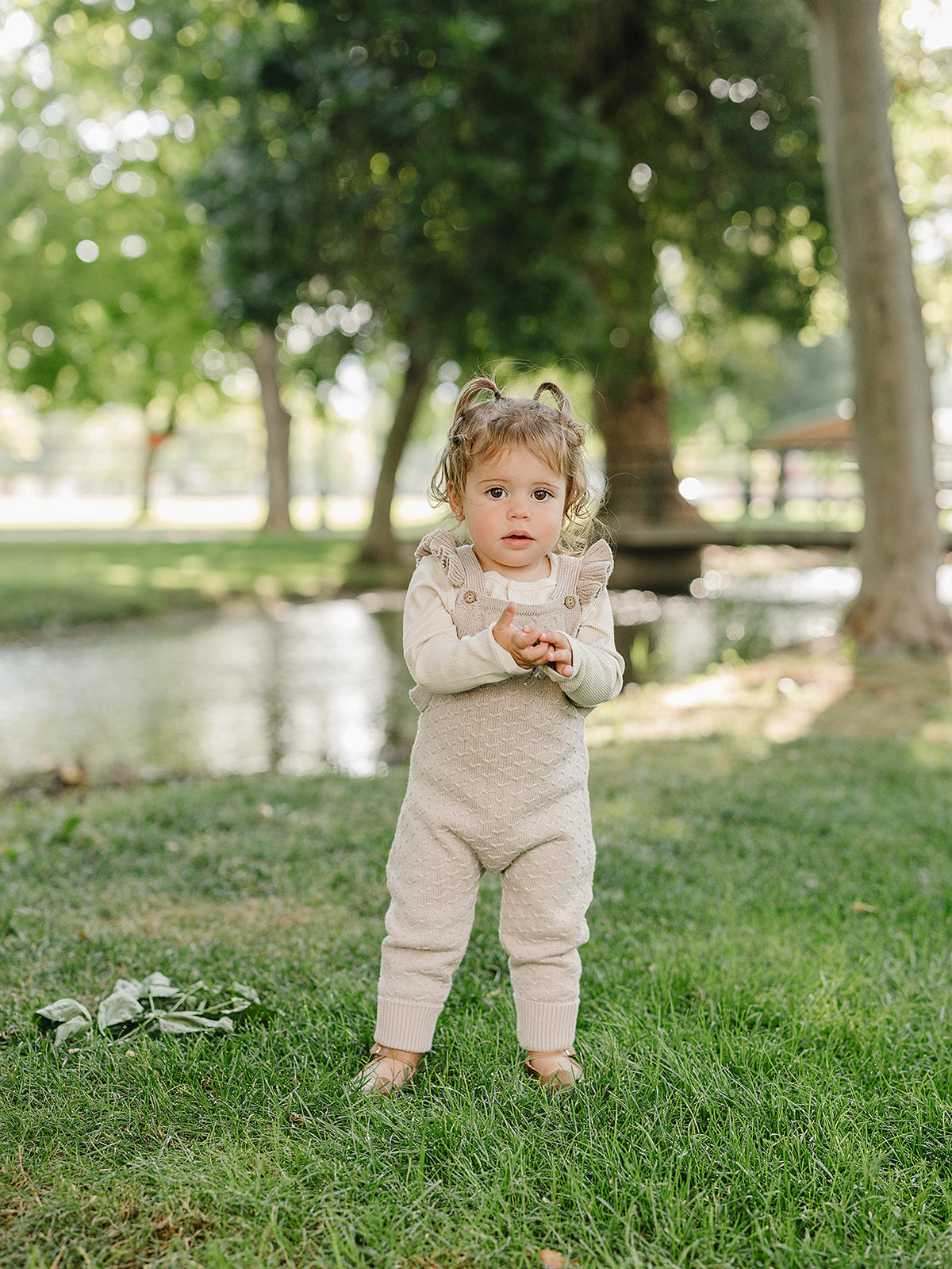 A toddler stands on green grass by a serene pond, wearing soft taupe knit overalls with ruffle details.