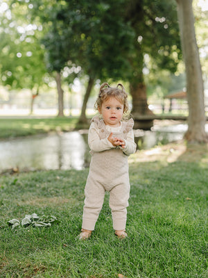 A toddler stands on green grass by a serene pond, wearing soft taupe knit overalls with ruffle details.