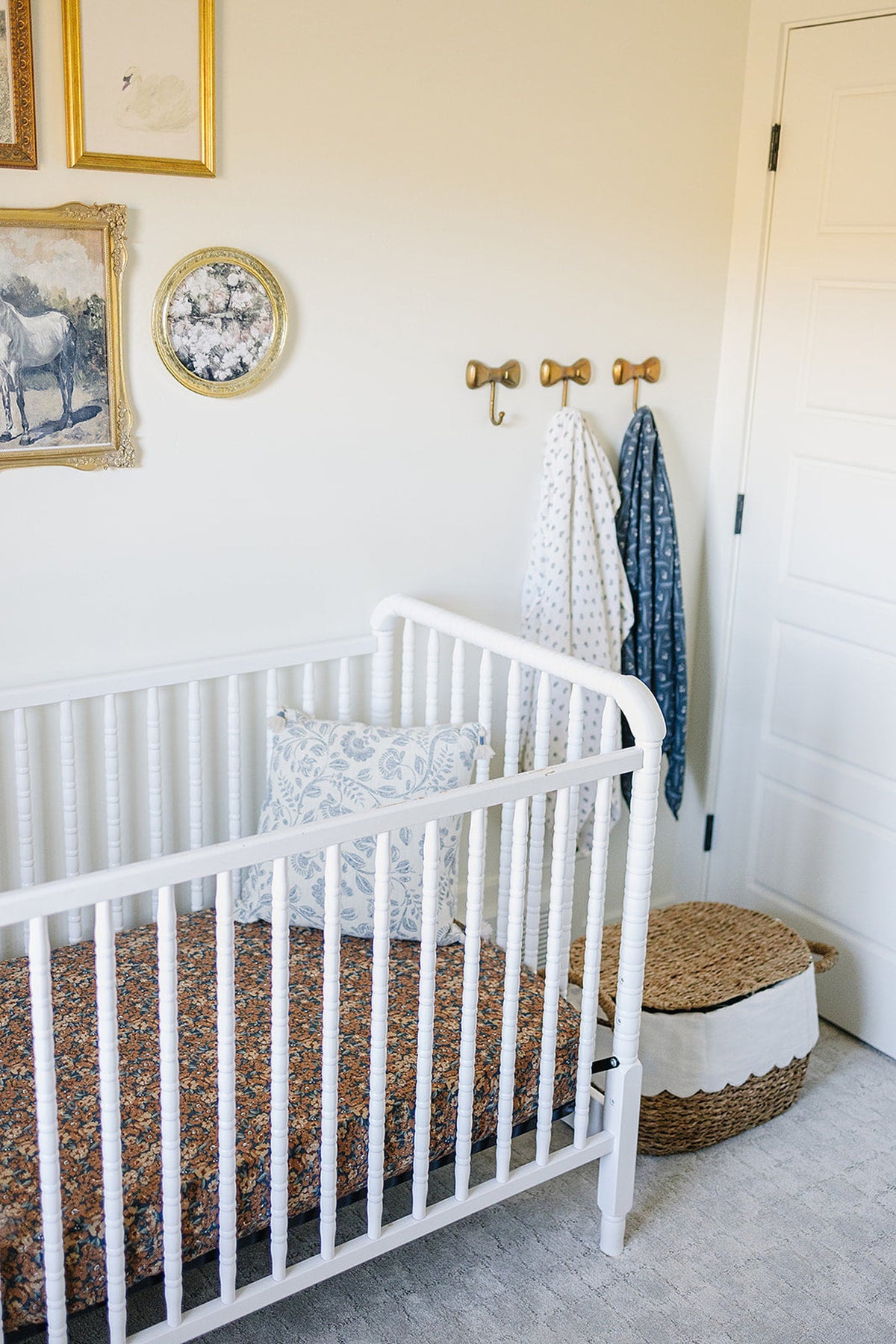 A white crib with a floral-patterned pillow and dark floral sheet, surrounded by decorative wall art and woven basket.