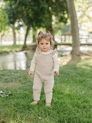 A toddler stands in a grassy area, wearing beige ruffle knit overalls and a long-sleeve cream top, surrounded by trees and a small body of water in the background.