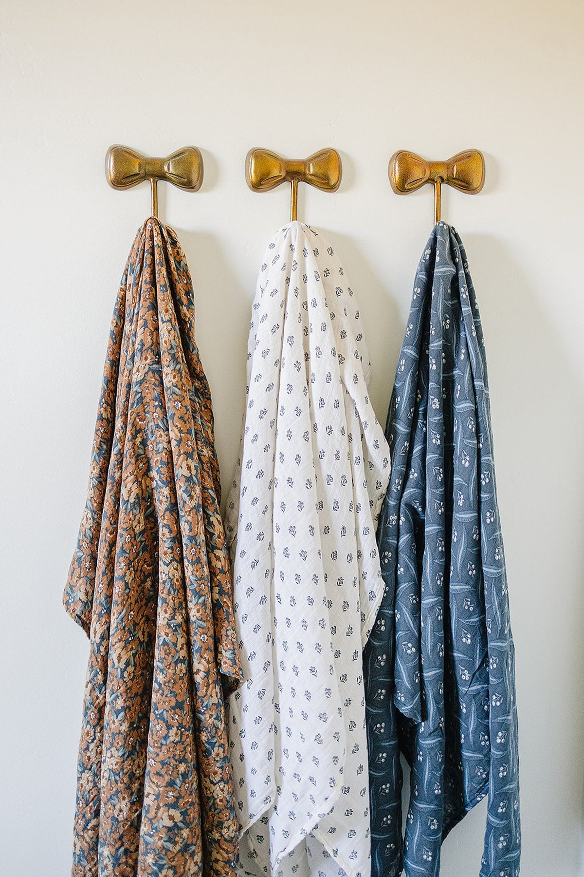 Three patterned muslin swaddle blankets hang on gold bow-shaped hooks: floral, white with small designs, and navy.