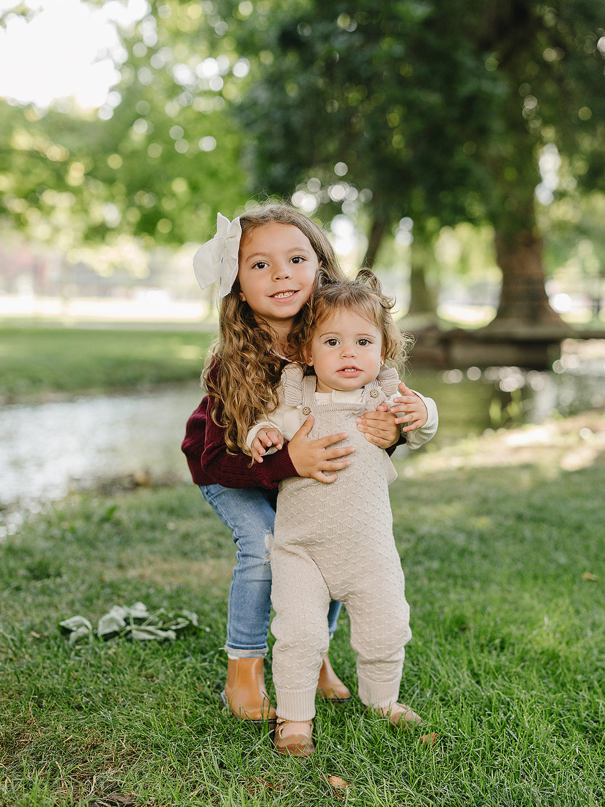 Two young girls stand in a grassy area near a serene pond, with one girl holding the other. They wear cozy, stylish outfits, showcasing a natural outdoor setting.