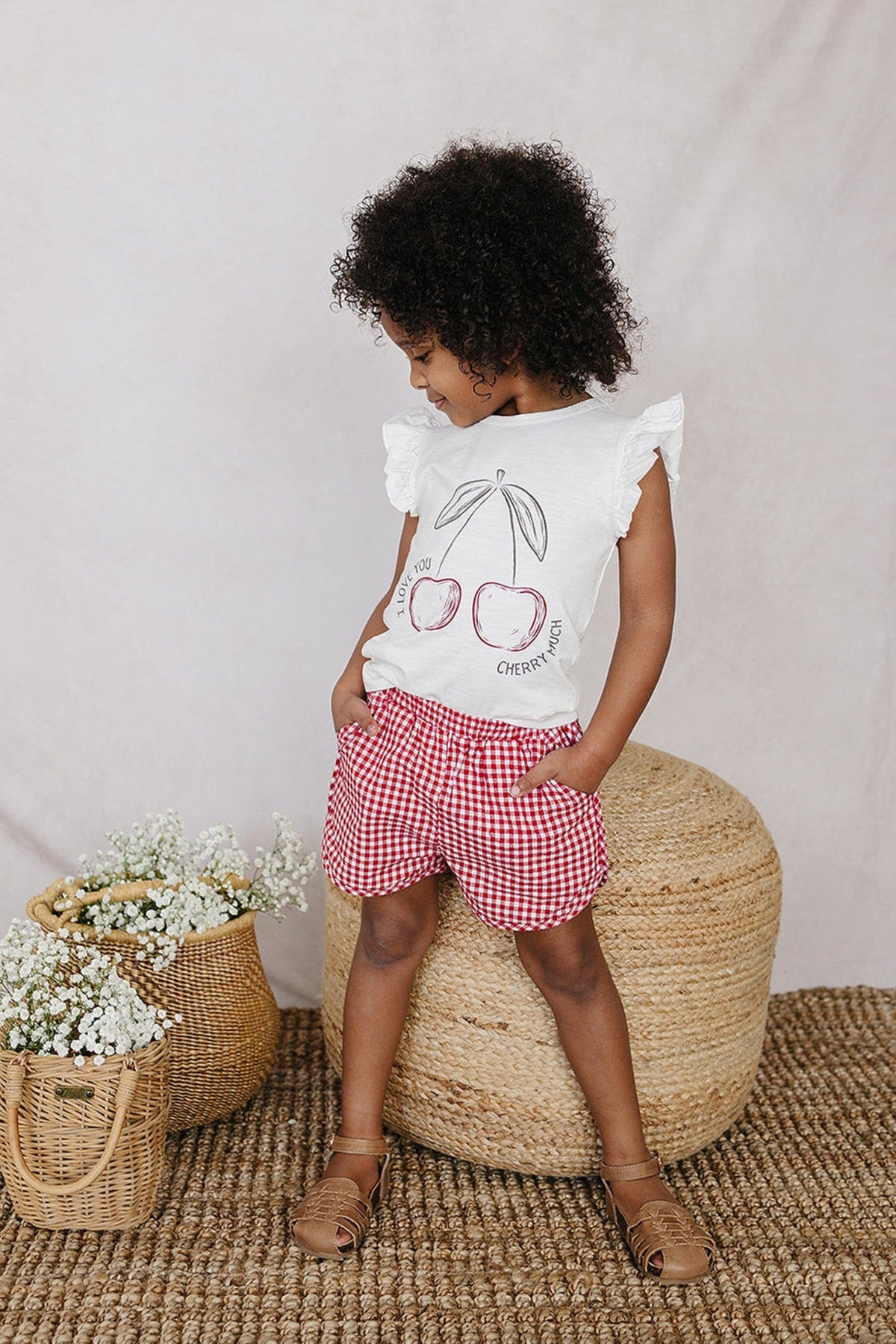 A child wearing red gingham shorts and a white cherry print top stands on a woven rug with baskets.