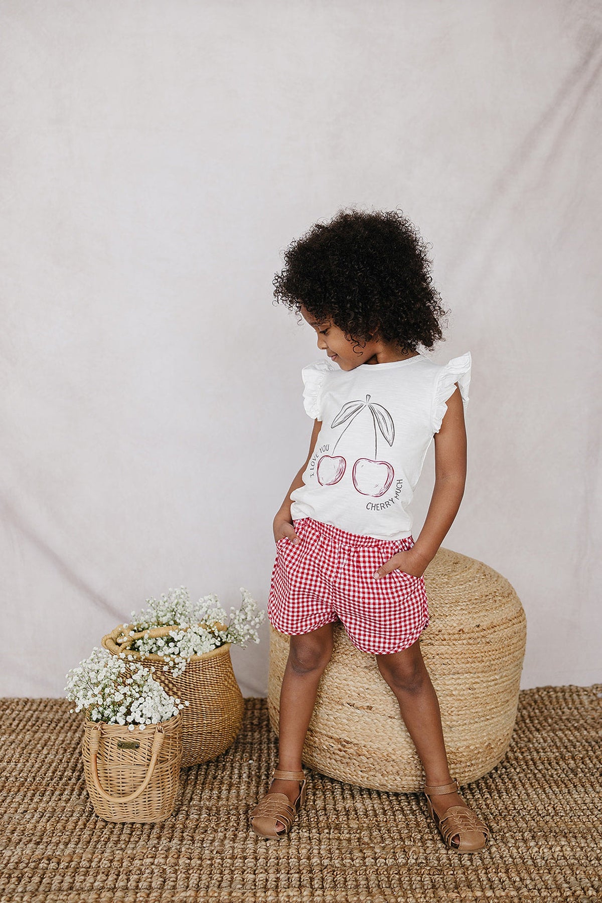 A child in a white cherry print ruffle T-shirt and red checkered shorts stands on a woven rug.
