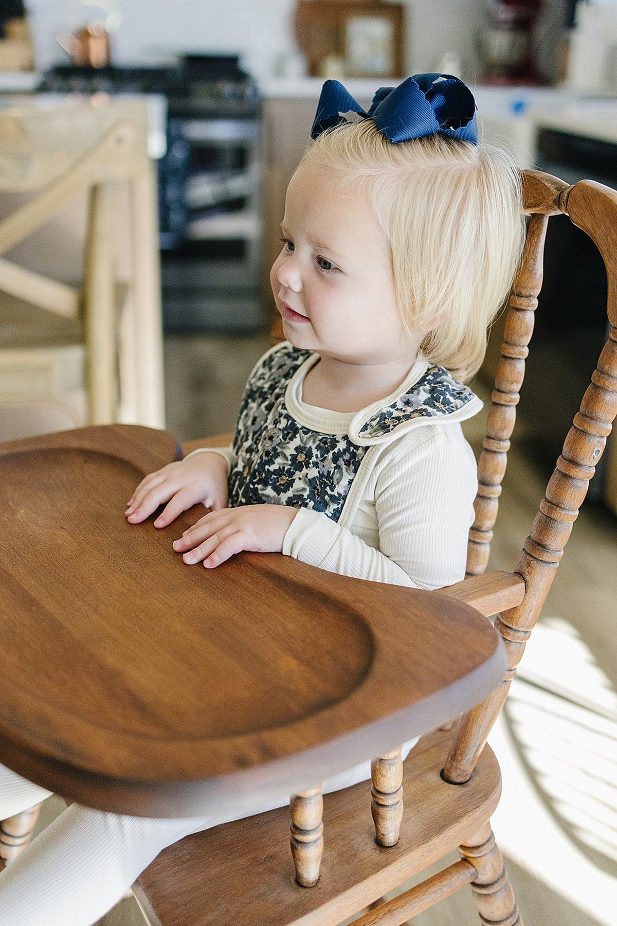 A child with a blue bow sits in a wooden high chair, wearing a floral-patterned bib and cream outfit.