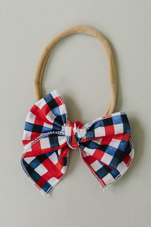 A handcrafted hair bow featuring a red, white, and blue plaid pattern is attached to a soft beige headband.