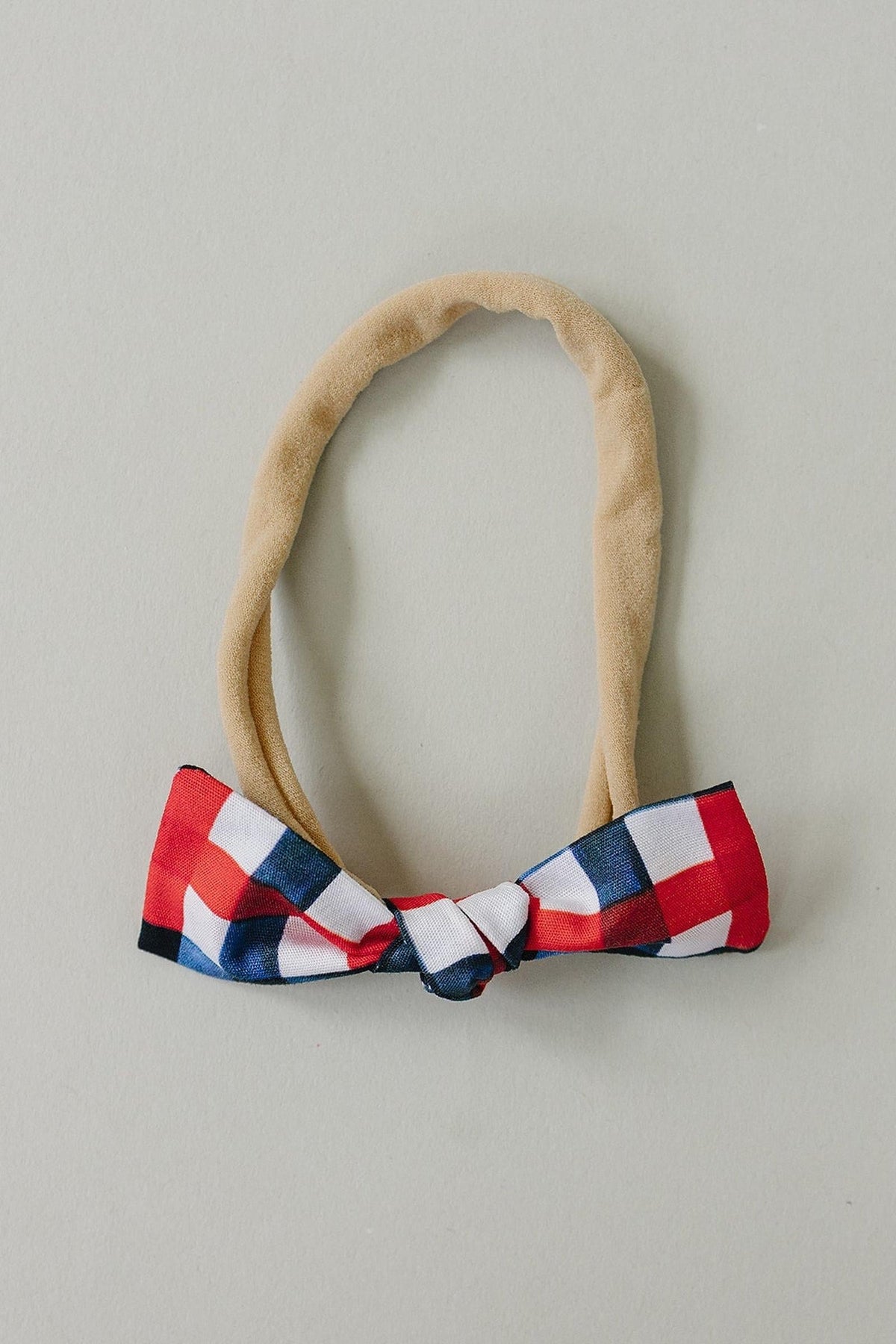 A handcrafted hair bow featuring a red, white, and blue plaid design, attached to a soft, beige elastic band. Ideal for children, this bow adds charm to any outfit.