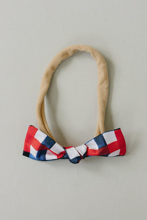 A handcrafted hair bow featuring a red, white, and blue plaid design, attached to a soft, beige elastic band. Ideal for children, this bow adds charm to any outfit.
