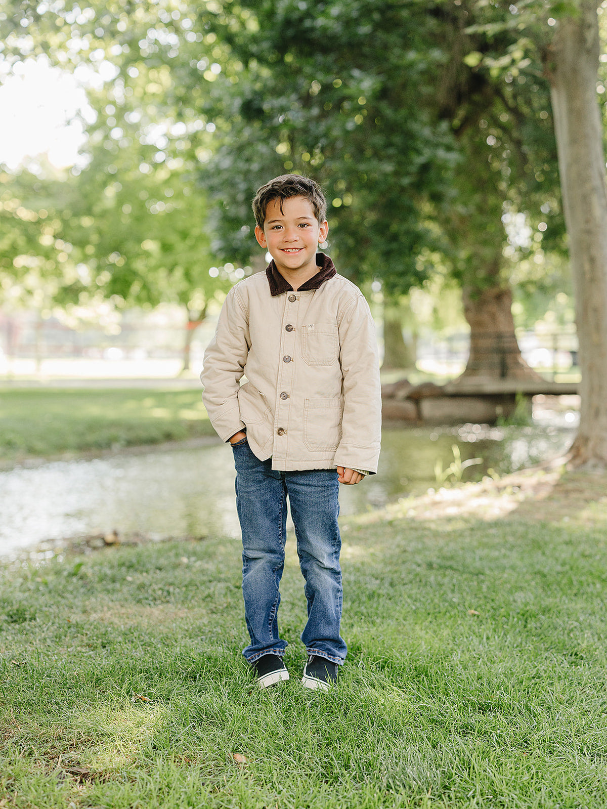 A young boy stands in a grassy area near a stream, wearing a light beige barn jacket over a brown collared shirt and blue jeans, showcasing a casual, stylish look.