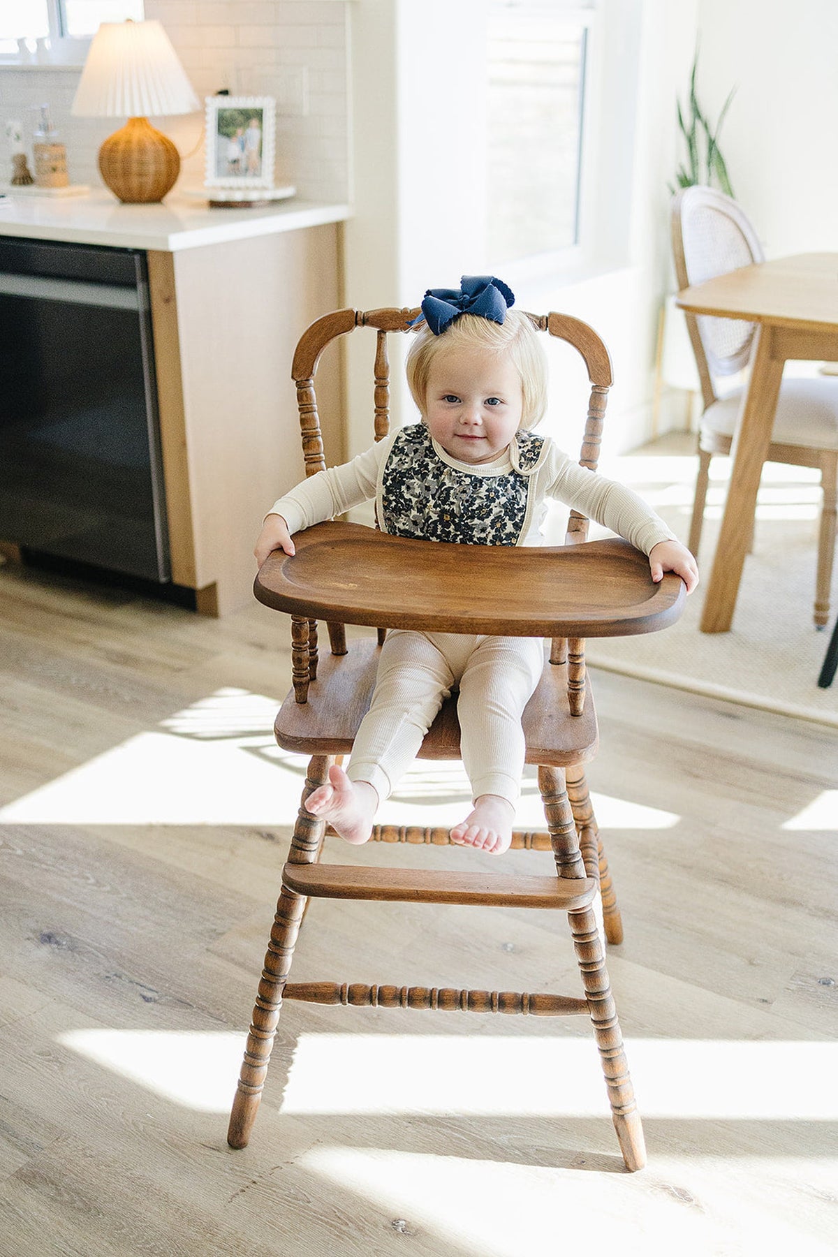 A baby wearing a floral bib and cream outfit sits in a wooden high chair in a sunlit kitchen.