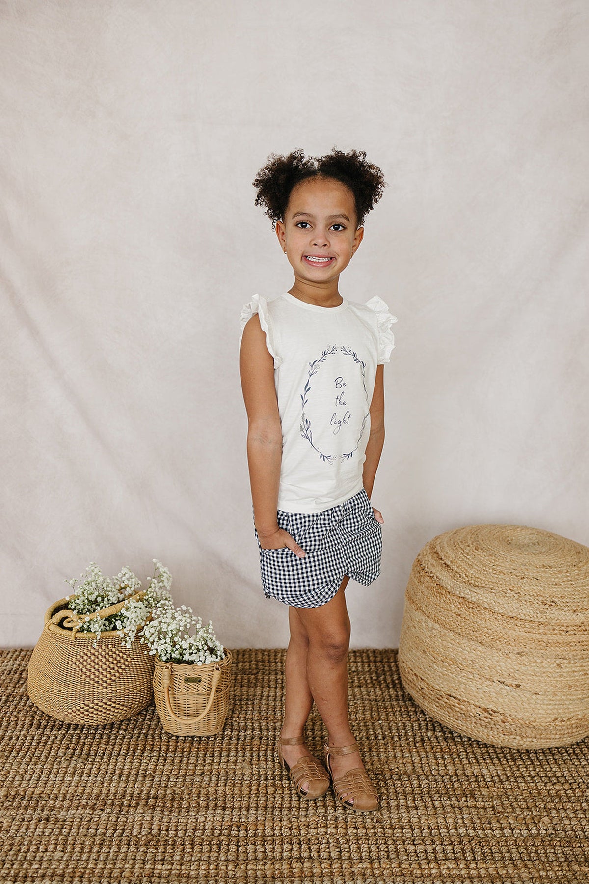 A child wearing a white ruffle-sleeve t-shirt with Be the Light text and checkered shorts stands smiling.