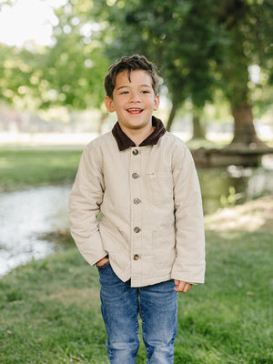 A smiling child stands outdoors by a stream, wearing a light beige cotton jacket with a brown collar, paired with blue jeans.