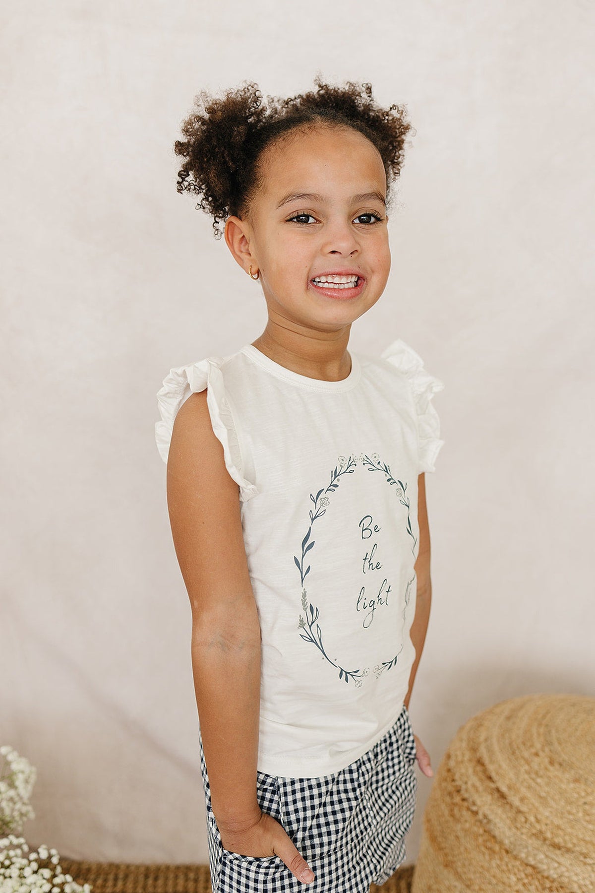 A smiling child wearing a white ruffle sleeve t-shirt with Be the light text, paired with checkered shorts.