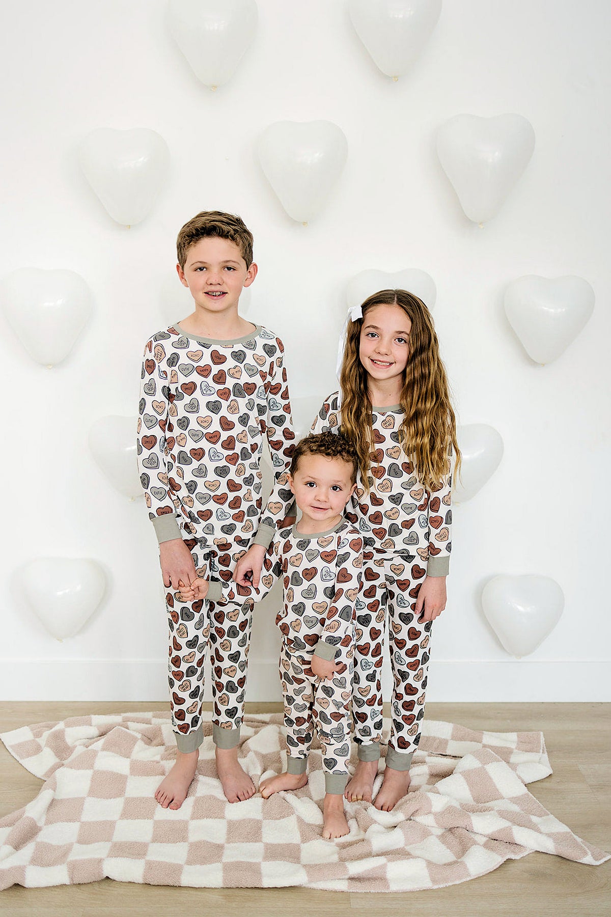 Three children wear matching ribbed pajamas with heart patterns, standing on a cozy blanket against a wall with heart-shaped balloons.