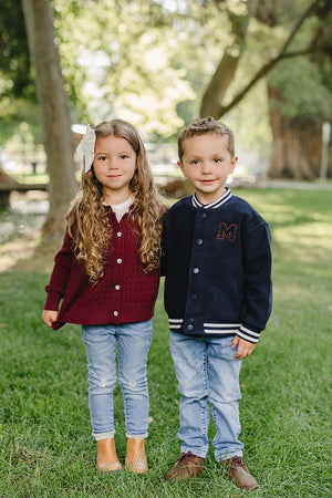 A girl with long curly hair wears a burgundy cable knit cardigan, while a boy in a navy varsity jacket stands beside her. They are outdoors on a grassy area.