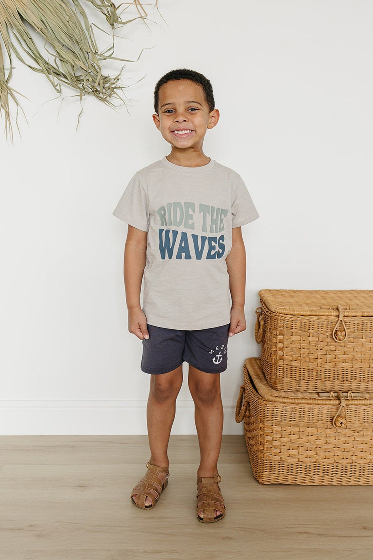 A smiling child wears a Ride the Waves t-shirt and dark shorts, standing near wicker baskets.