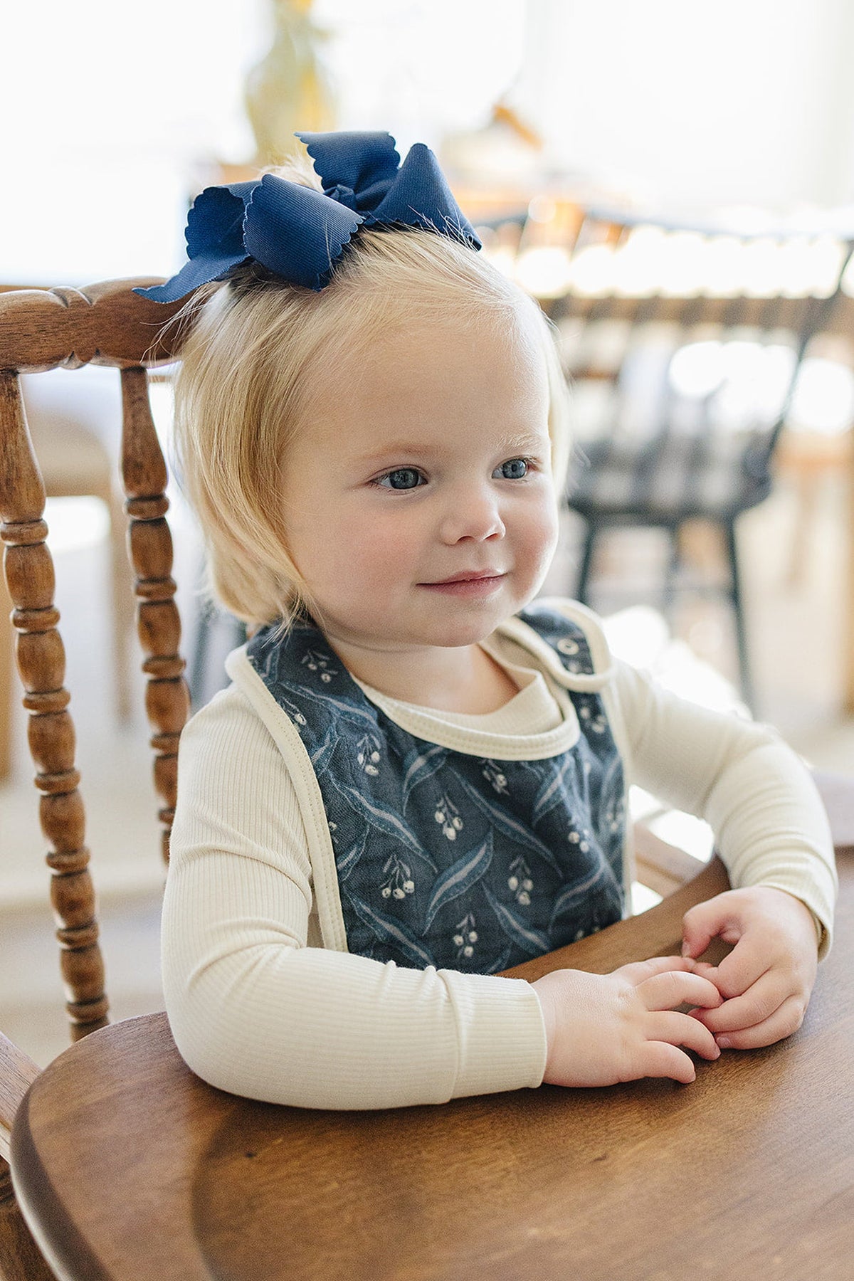 A child wearing a navy patterned bib and cream top sits in a wooden chair with a blue bow.