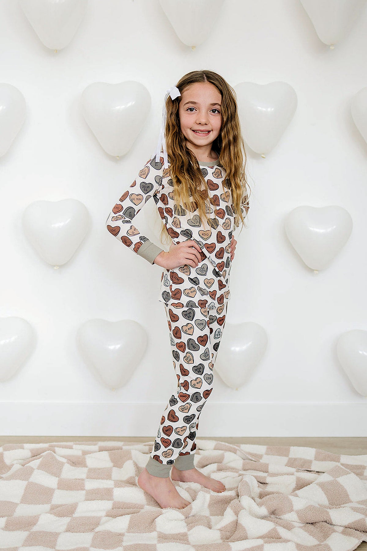 A smiling girl poses in a cozy ribbed pajama set featuring heart patterns, with white balloons in the background.