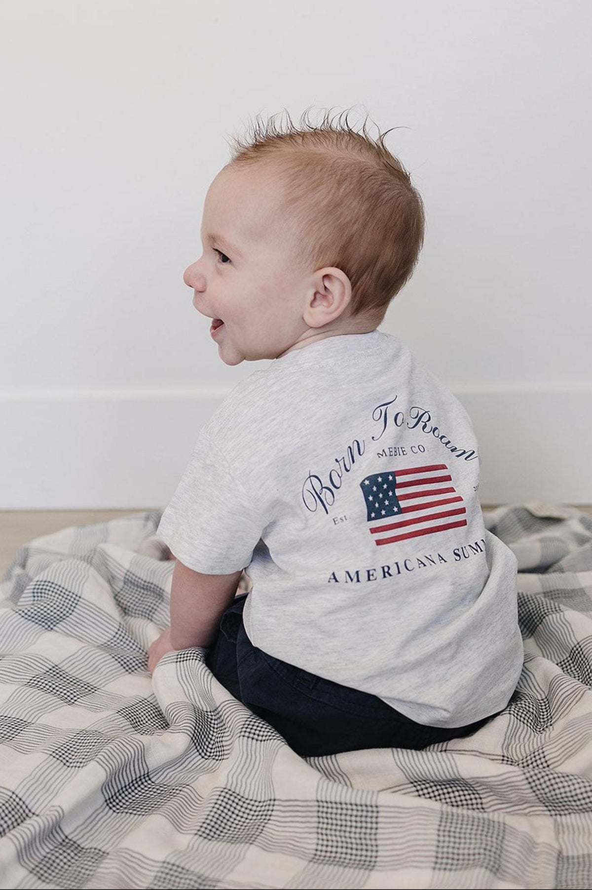 A baby wearing a gray Mebie Baby T-shirt with an American flag graphic sits on a checkered blanket.