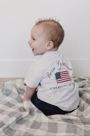 A baby wearing a gray Mebie Baby T-shirt with an American flag graphic sits on a checkered blanket.